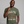 A smiling man wearing an olive green graphic t-shirt with a skeleton dancing among red and orange autumn leaves. The text on the shirt reads 'IT'S THE MOST WONDERFUL TIME OF THE YEAR'.