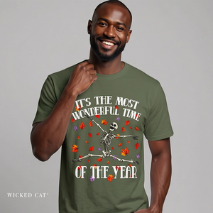 A smiling man wearing an olive green graphic t-shirt with a skeleton dancing among red and orange autumn leaves. The text on the shirt reads 'IT'S THE MOST WONDERFUL TIME OF THE YEAR'.