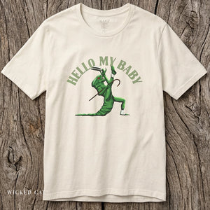 An off-white crewneck short-sleeve crewneck t-shirt featuring a green alien dancing, with the text 'HELLO MY BABY' arched above it, displayed flat on a rustic wood background.