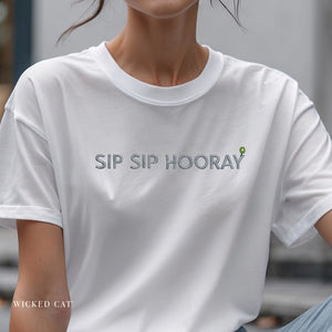 White t-shirt with 'SIP SIP HOORAY' text and a green leaf design on a blurred background