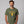 A man wearing an olive green t-shirt with a yellow and blue graphic of a soldier and the text 'FUSILLI JERRY' on the front, set against a solid gray background.