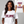 A woman wearing a white graphic T-shirt with the phrase 'START JUMPING' in a dark burgundy stylized font across the chest. A smaller inset shows the back of the same shirt with the phrase 'SHAKE THE STADIUM' in matching burgundy and orange lettering.