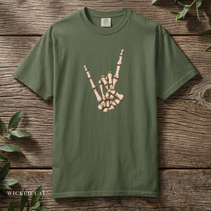 An olive green short-sleeve t-shirt with a graphic of a skeleton hand making the rock on gesture, displayed flat on a rustic wooden background with green leaves.