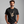 A man wearing a black crewneck t-shirt with a beige skeleton hand graphic making a rock on gesture, set against a plain gray studio background. The brand name 'WICKED CAT' is printed in white in the bottom-left corner.