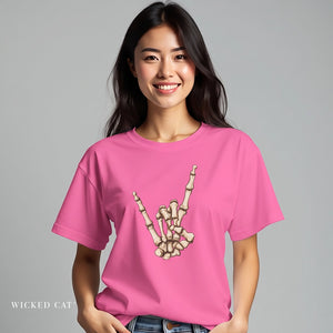 A woman wearing a bright pink t-shirt with a graphic of a skeleton hand making a rock on sign, set against a solid gray background. The brand name 'WICKED CAT' is visible in the bottom left corner.
