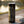 A tall black insulated tumbler with a clear lid and metal straw sits on a stone ledge outdoors near a blurred body of water. The tumbler has gothic-style printed artwork and text, including 'Book of Shadows' and 'Wicked Cat.
