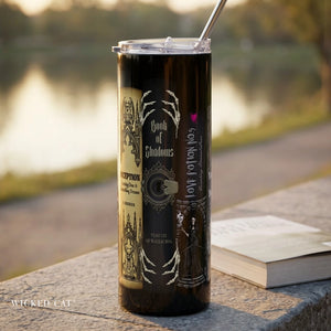 A tall black insulated tumbler with a clear lid and metal straw sits on a stone ledge outdoors near a blurred body of water. The tumbler has gothic-style printed artwork and text, including 'Book of Shadows' and 'Wicked Cat.