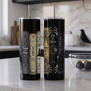Two tall black skinny tumblers with clear lids and metal straws, standing on a kitchen counter. The left tumbler features gothic book spine artwork, and the right tumbler features gothic book cover artwork with the text 'Book of Shadows' and 'Wicked Cat'.